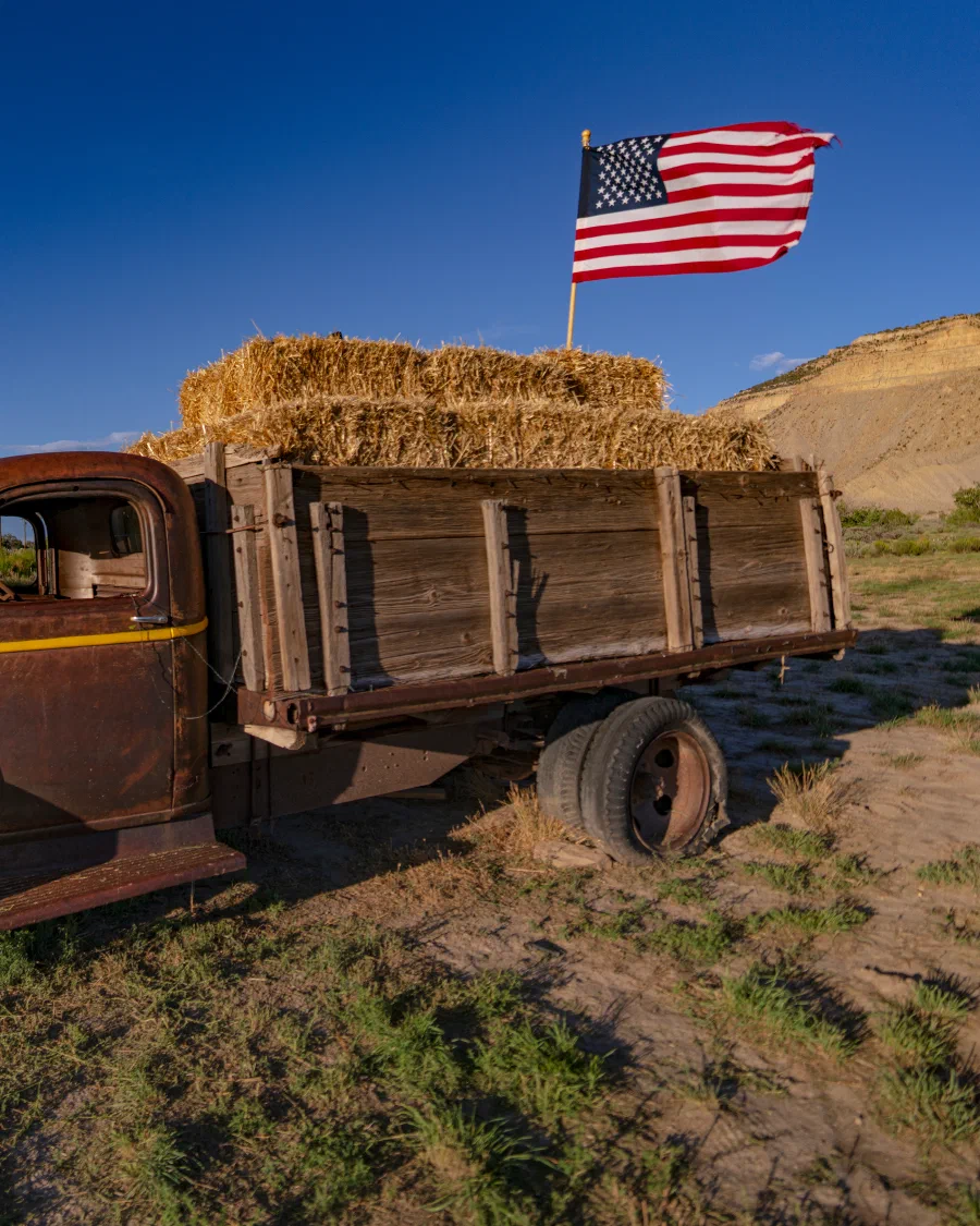 A vintage farm truck loaded with hay bales beneath an American flag in the Colorado high desert